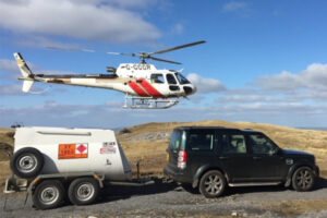 A white helicopter hovers low over the moors beside a four-by-four towing a Fuel Proof Aviation Fuel Bowser